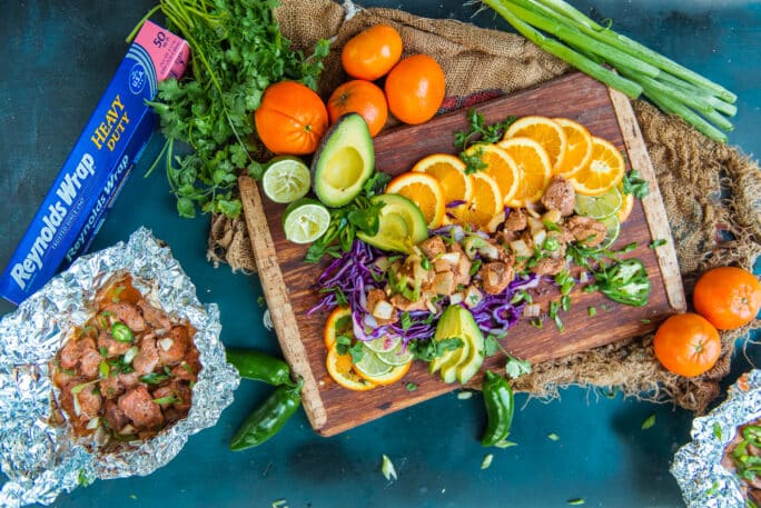 Flat lay of pork tenderloin bites over fresh sliced citrus with various fruit and cabbage garnish on cutting board and blue background.