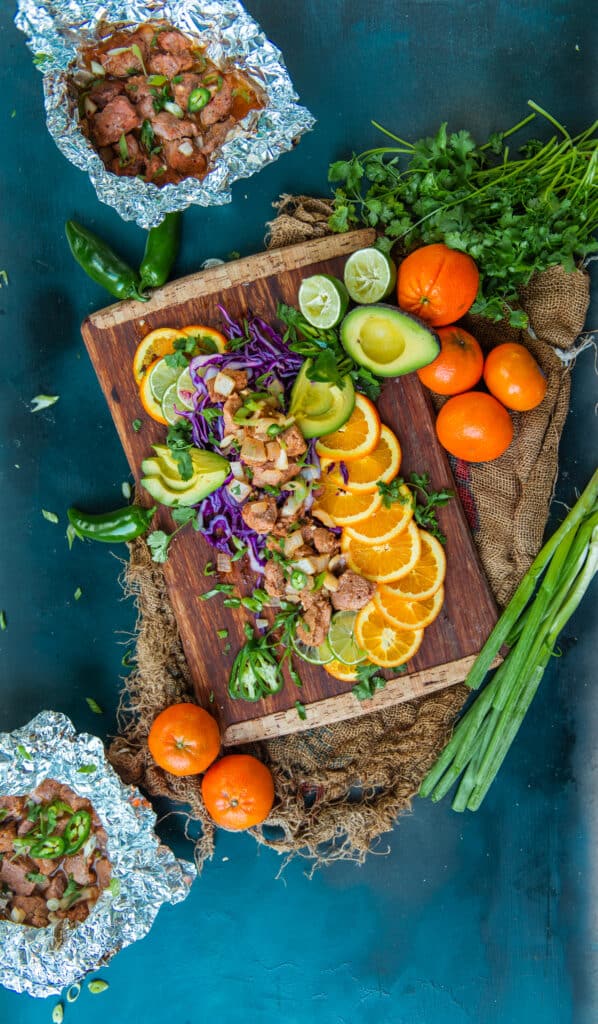 Pork tenderloin bites and shown in foil packets for ease of cooking on a blue background and cutting board with fresh citrus and other garnish