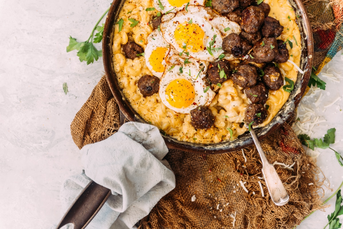 skillet full of creamy mac and cheese, topped with meatballs and fried eggs on white background