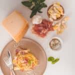 Flat lay ingredients for classic carbonara with a plate of finished pasta swirled with prosciutto and shaved cheese