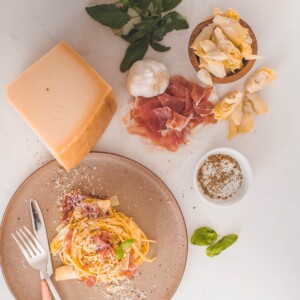 Flat lay ingredients for classic carbonara with a plate of finished pasta swirled with prosciutto and shaved cheese