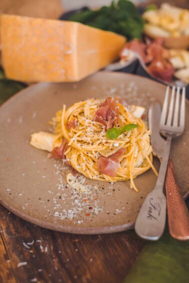 close up of a grey plate of twirled pasta carbonara, speckled with basil and prosciutto and topped with shaved cheese