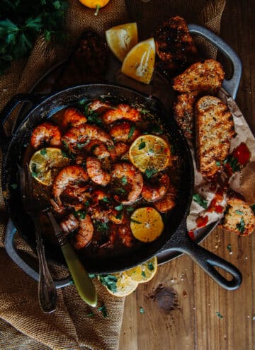 cast iron skillet filled with spicy sauce, jumbo shrimp, lemons and bread on the side for dipping