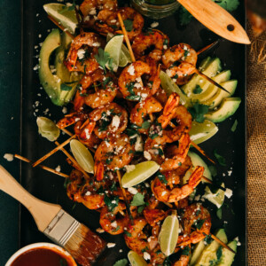 platter of bbq shrimp skewers grilled served with bbq sauce, over avocado slices, garnished with lime wedges, cotija and chimichurri