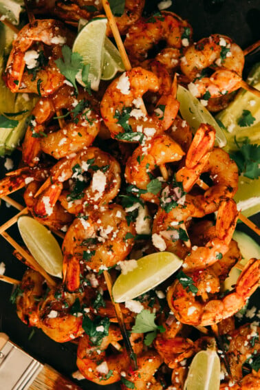 Bbq shrimp skewers on platter with crumbled cotija, brushed with chimichurri and lime wedges as garnish