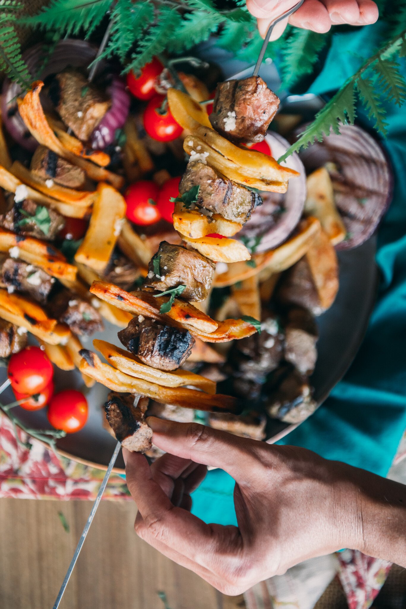 Beef and potato kabobs on platter with grilled onions and tomatoes with hands grabbing skewer.