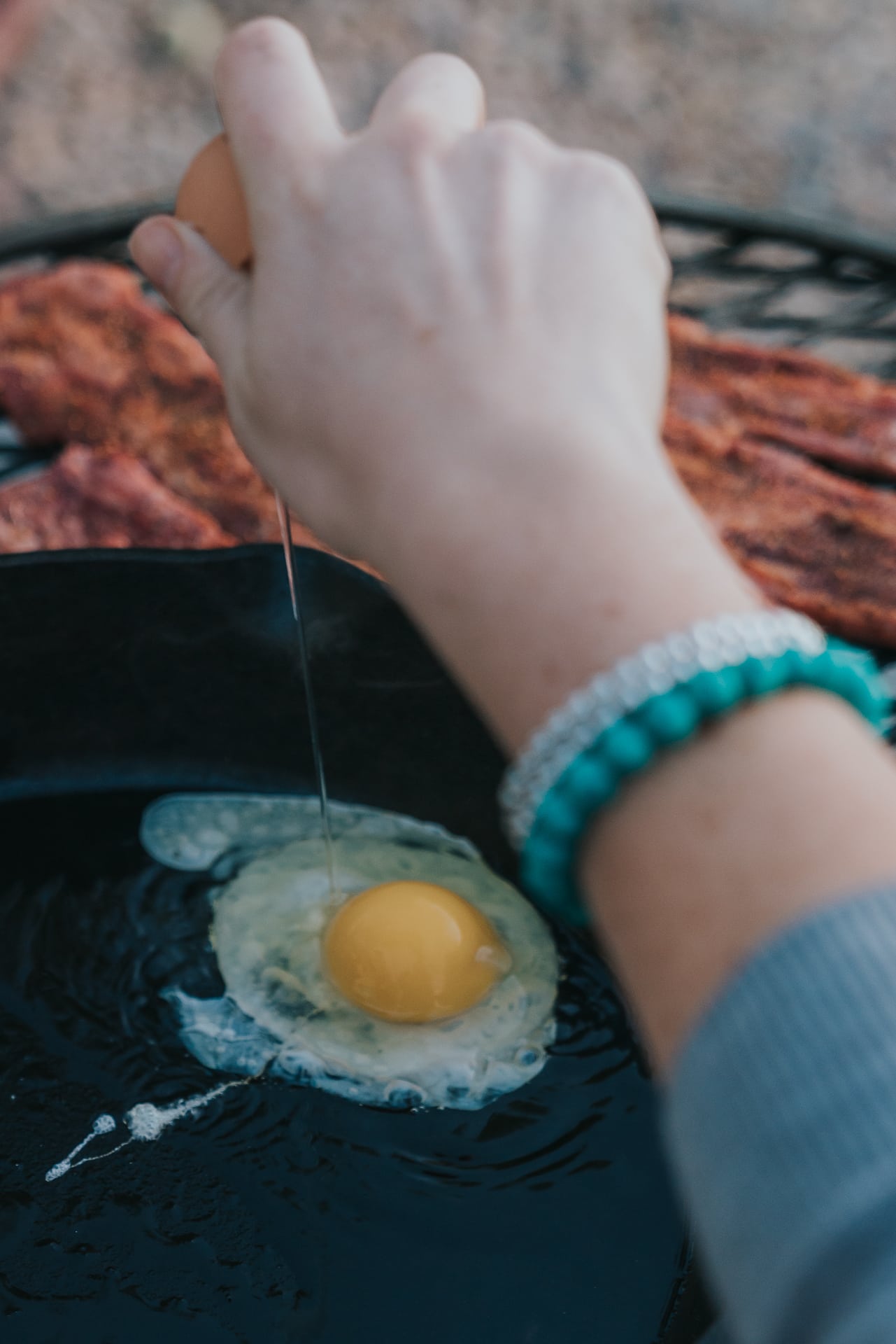 Hand cracking an egg into a cast iron skillet over campfire.