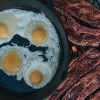 short ribs on a grilling grate over coals with a cast iron skilled filled with eggs