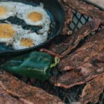 fire grate over the fire filled with cast iron filled with fried eggs, grilled beef, and peppers