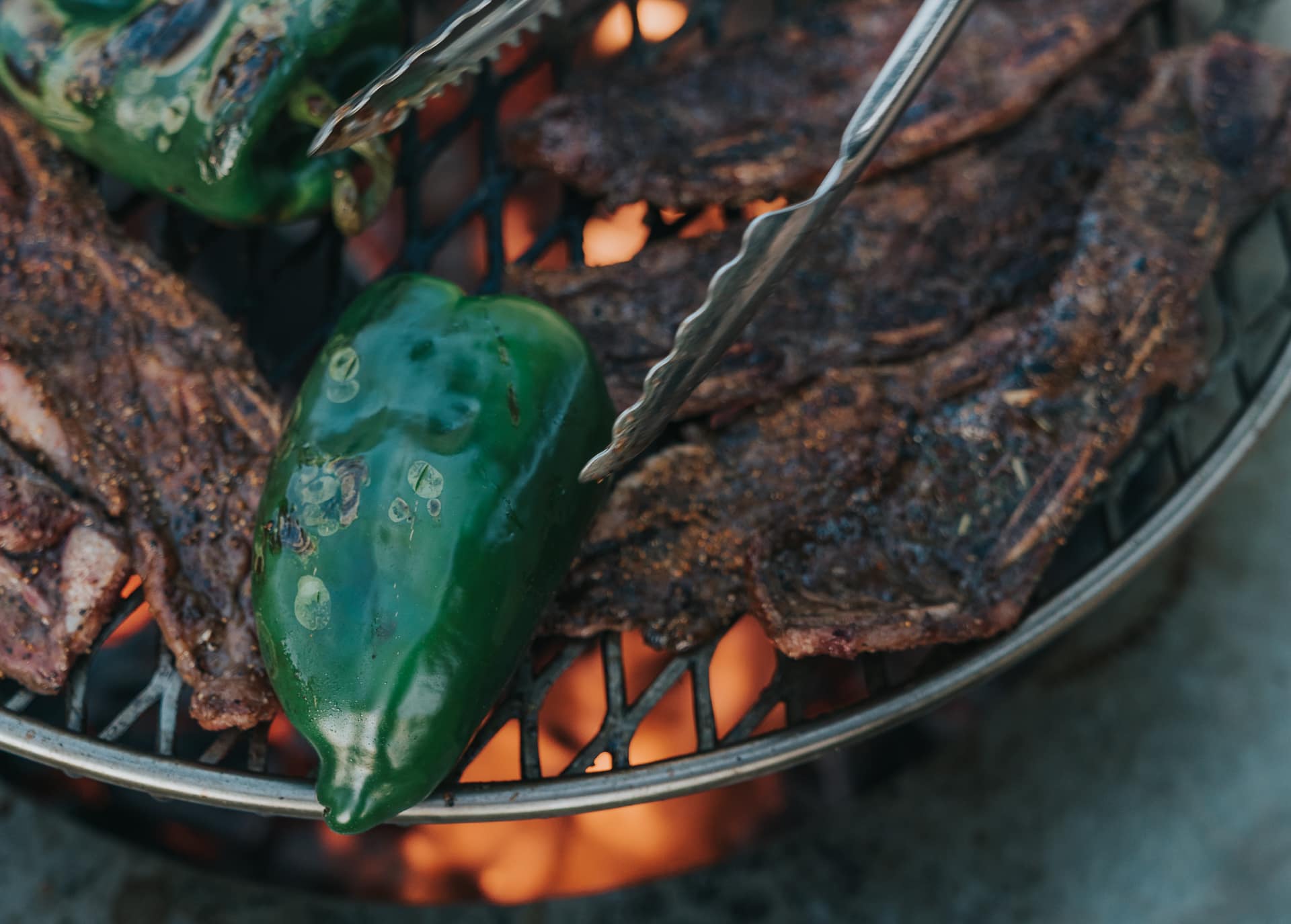 Poblano peppers and beef ribs on a grill grate over flames. 