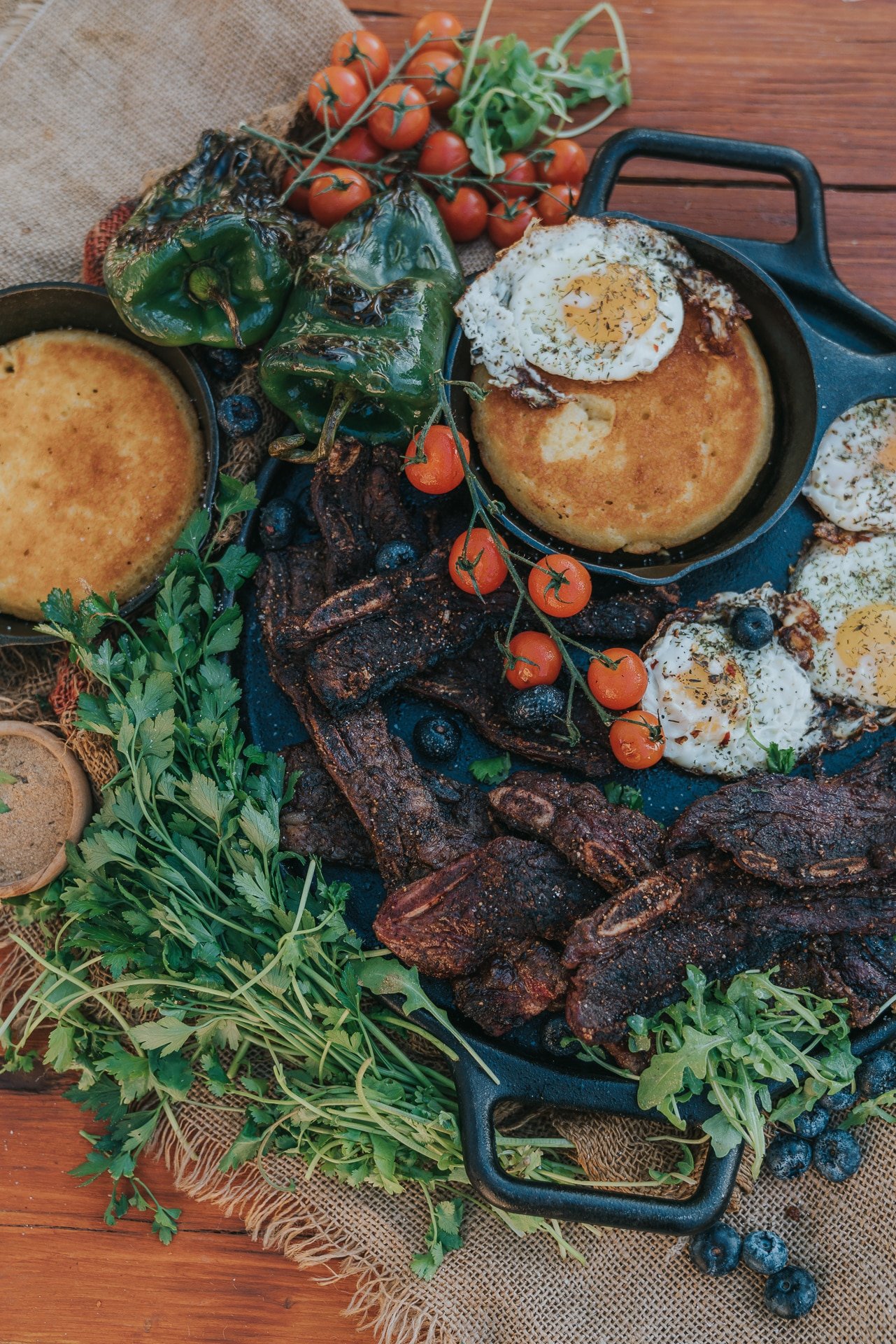 Overhead platter of short ribs, fried eggs, tomatoes and more in a rustic camp scene. 