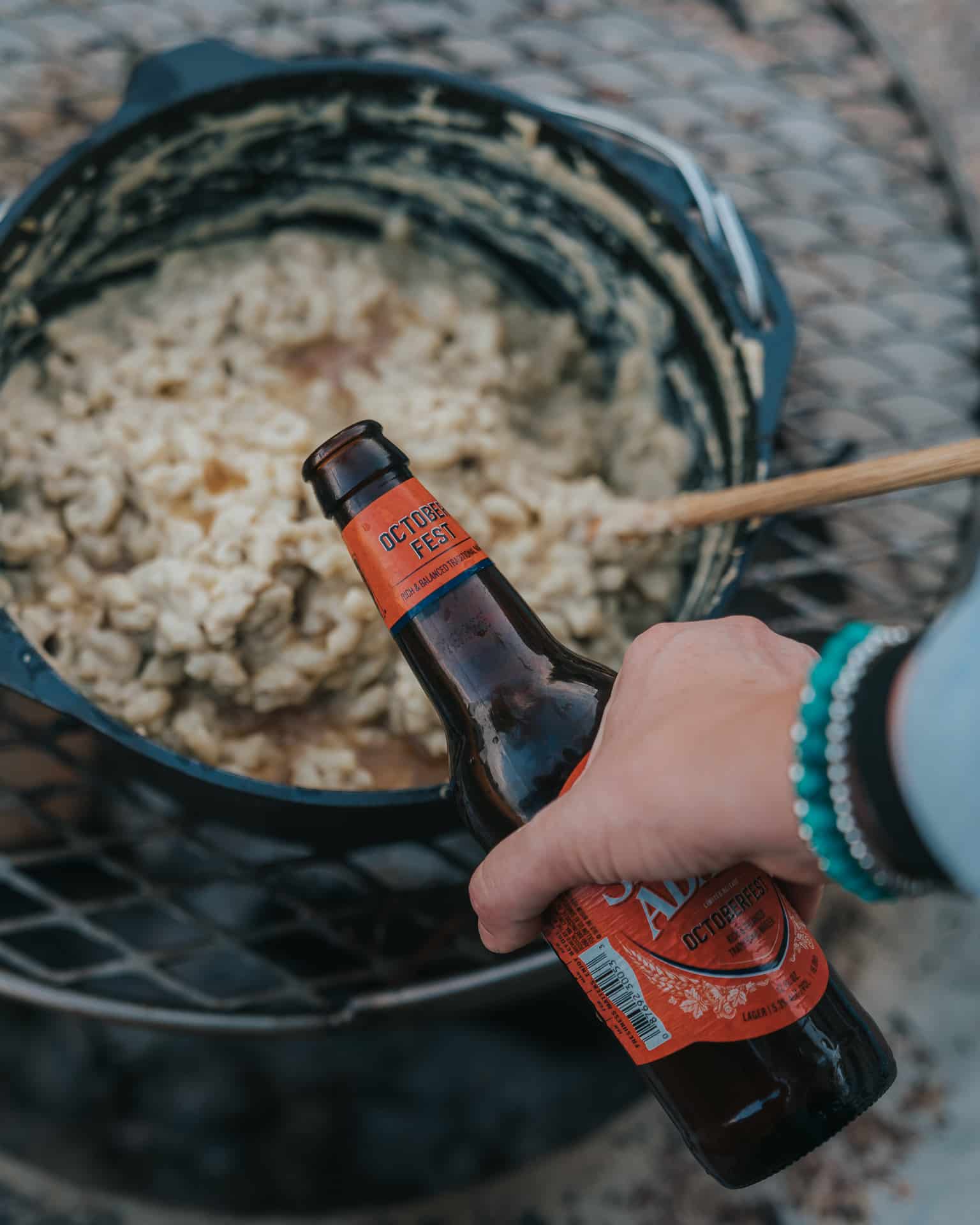 Hand pouring beer into cast iron pot of mac and cheese. 