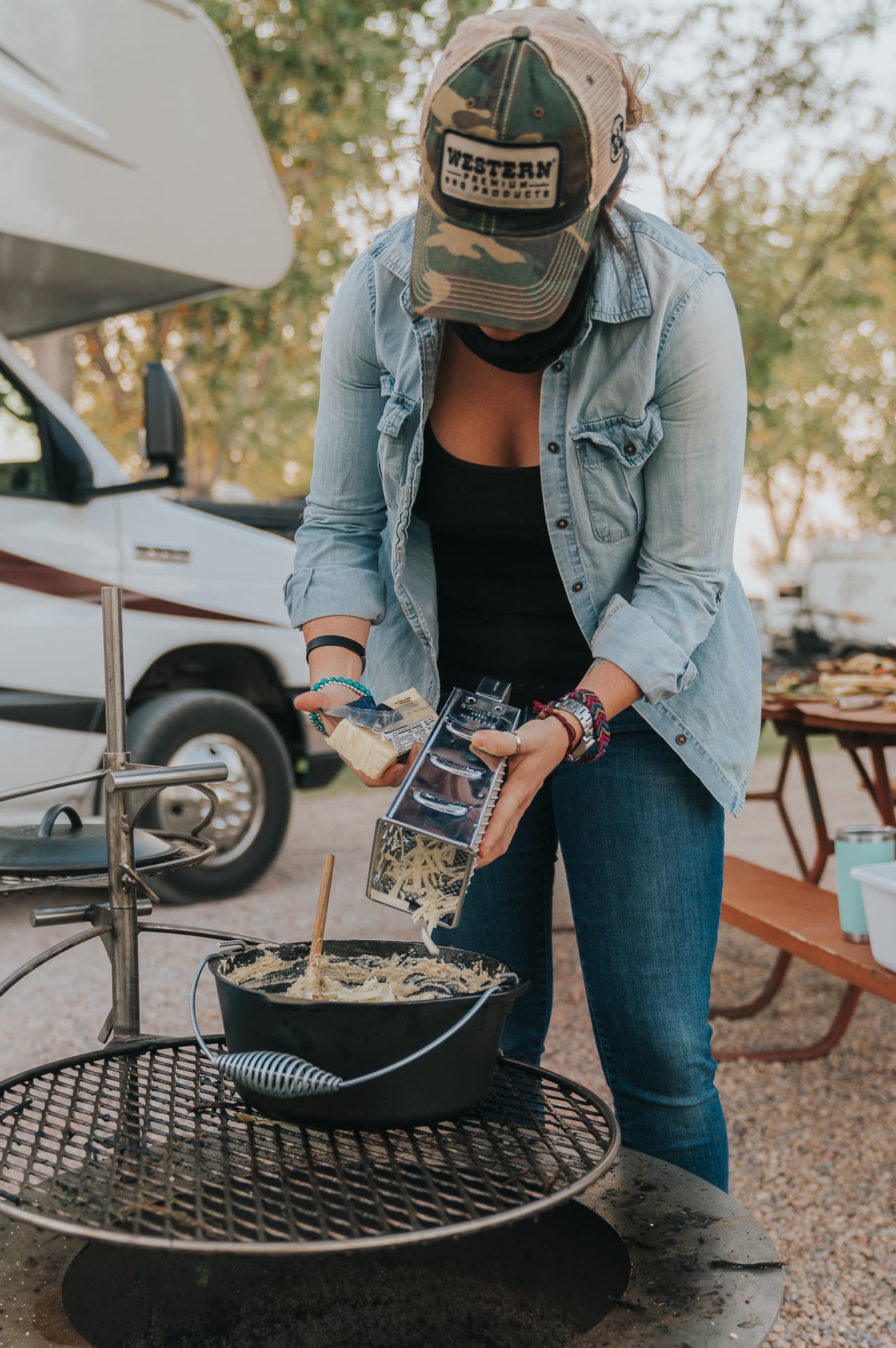 Woman grating cheese into a dutch oven over a fire pit. 