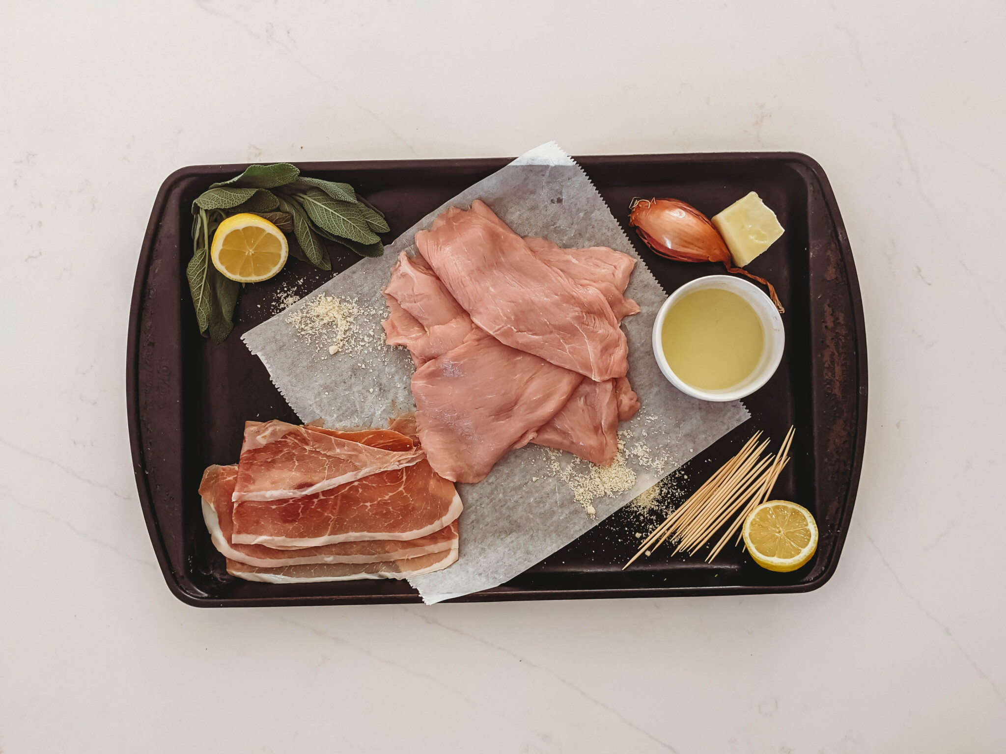 ingredients for veal saltimbocca; raw veal cutlets, lemon, prosciutto, sage, butter, oil