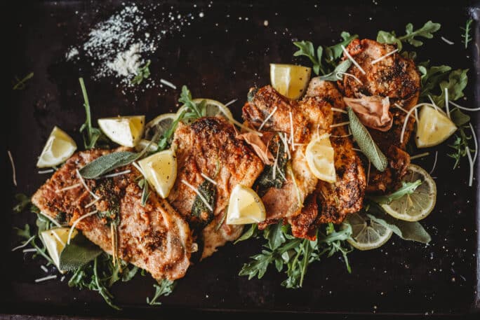 cooked veal saltimbocca on dark platter with fresh green arugula and lemon wedges