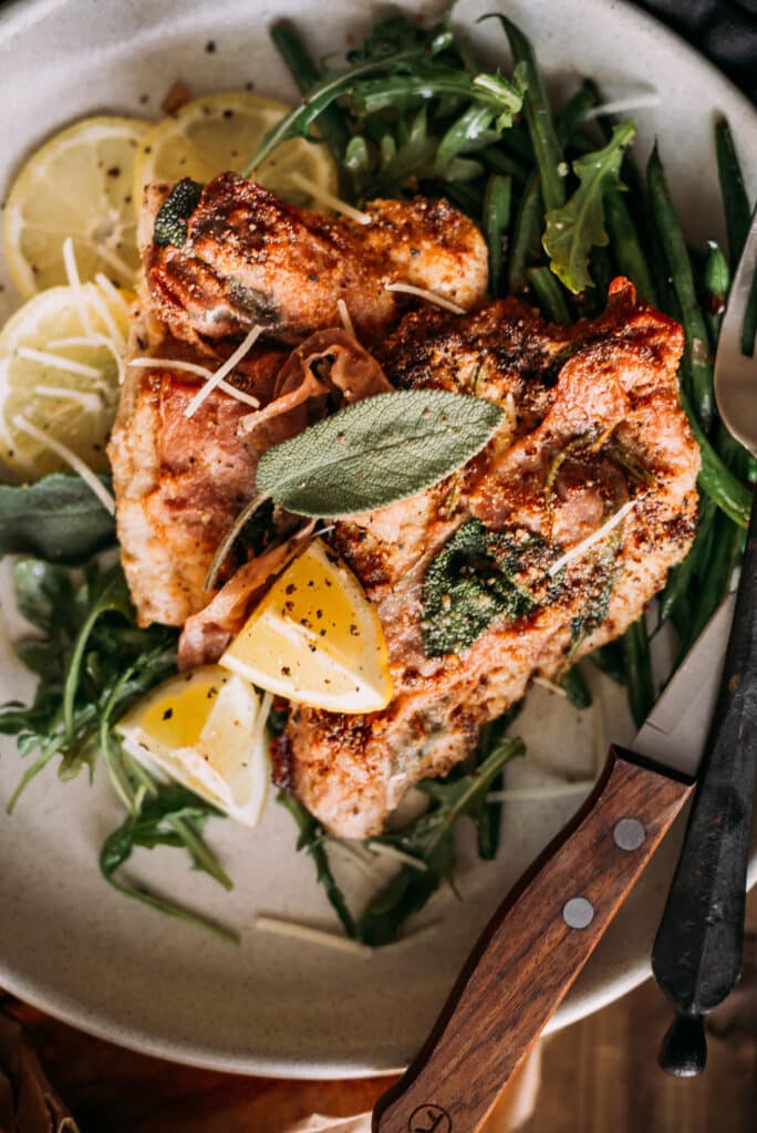 Cooked veal saltimbocca on a platter with fresh sage leaf, lemon wedges and arugula