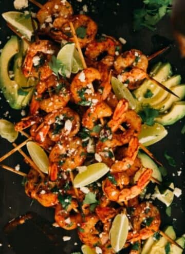 BBQ shrimp skewers on a platter with lime wedges, avocado slices, and cotija cheese.