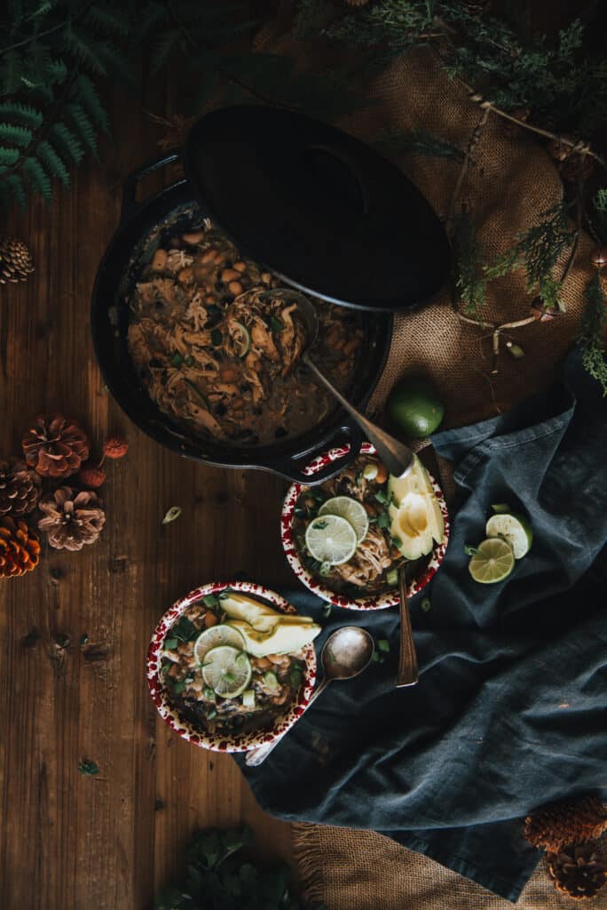 Above shot showing white chicken chili in a dutch oven with two bowls assembled near by topped with avocado and lime