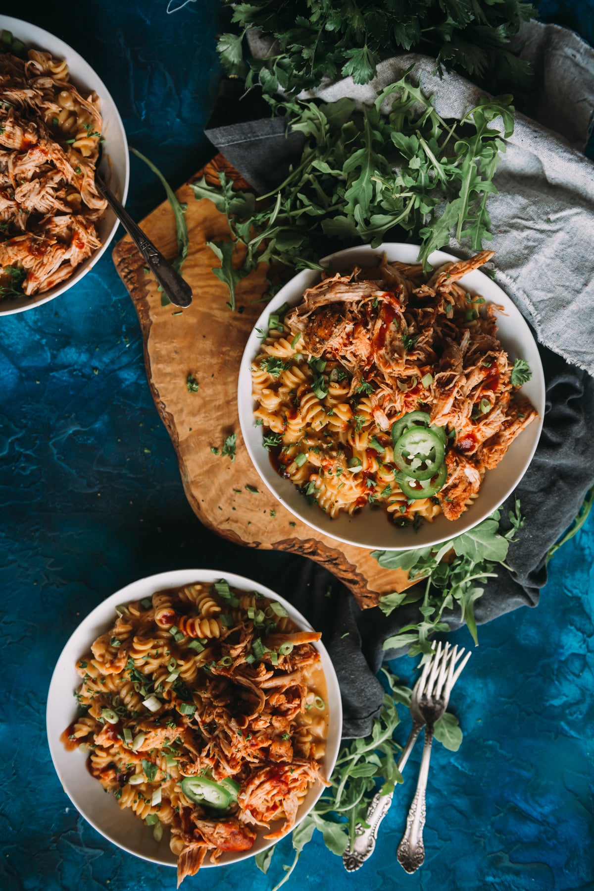 3 bowls in instant pot bbq chicken mac and cheese on a blue background topped with fresh herbs, jalapenos and extra bbq sauce