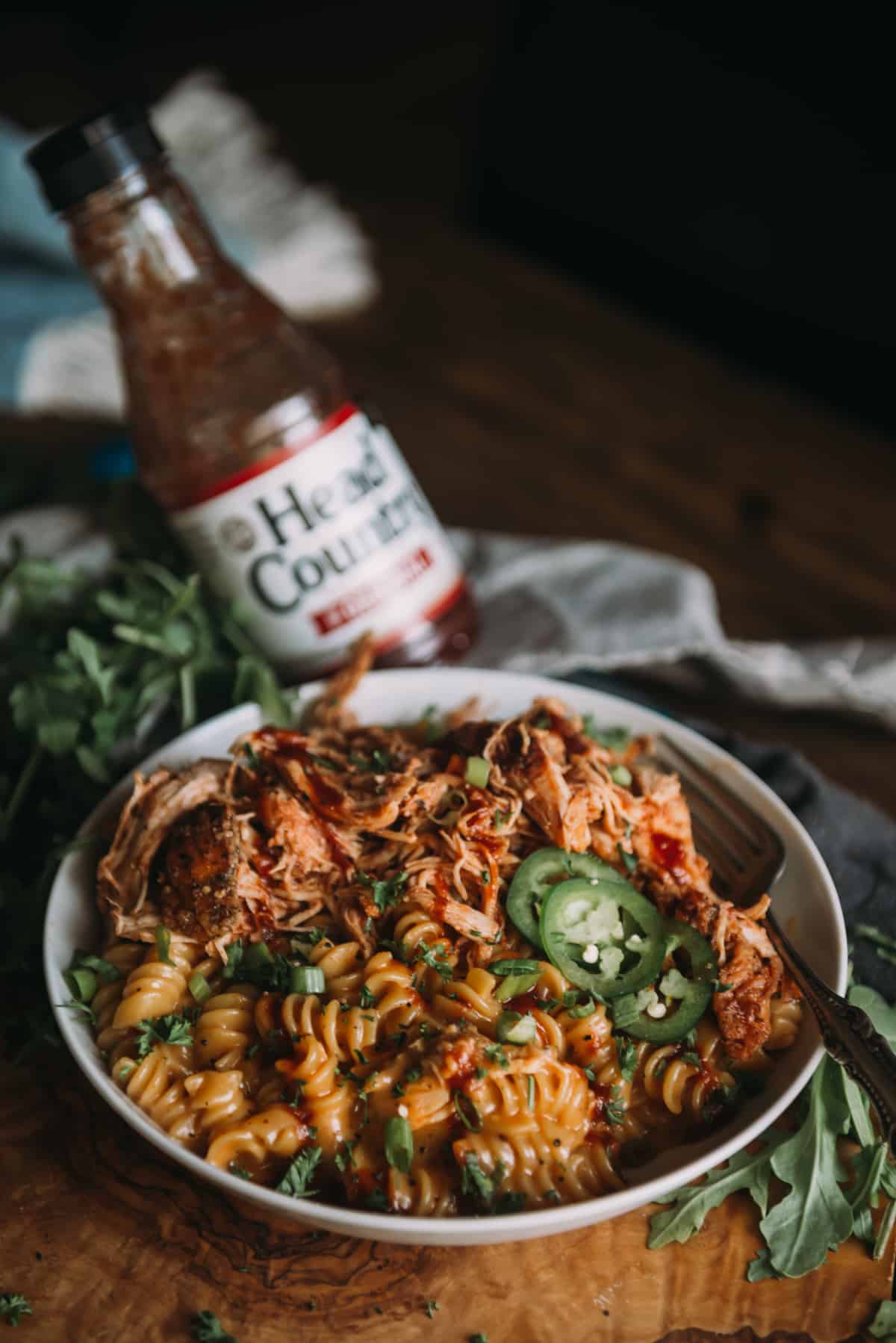 bowl of creamy shredded bbq chicken mac and cheese with bottle of head country bbq sauce behind