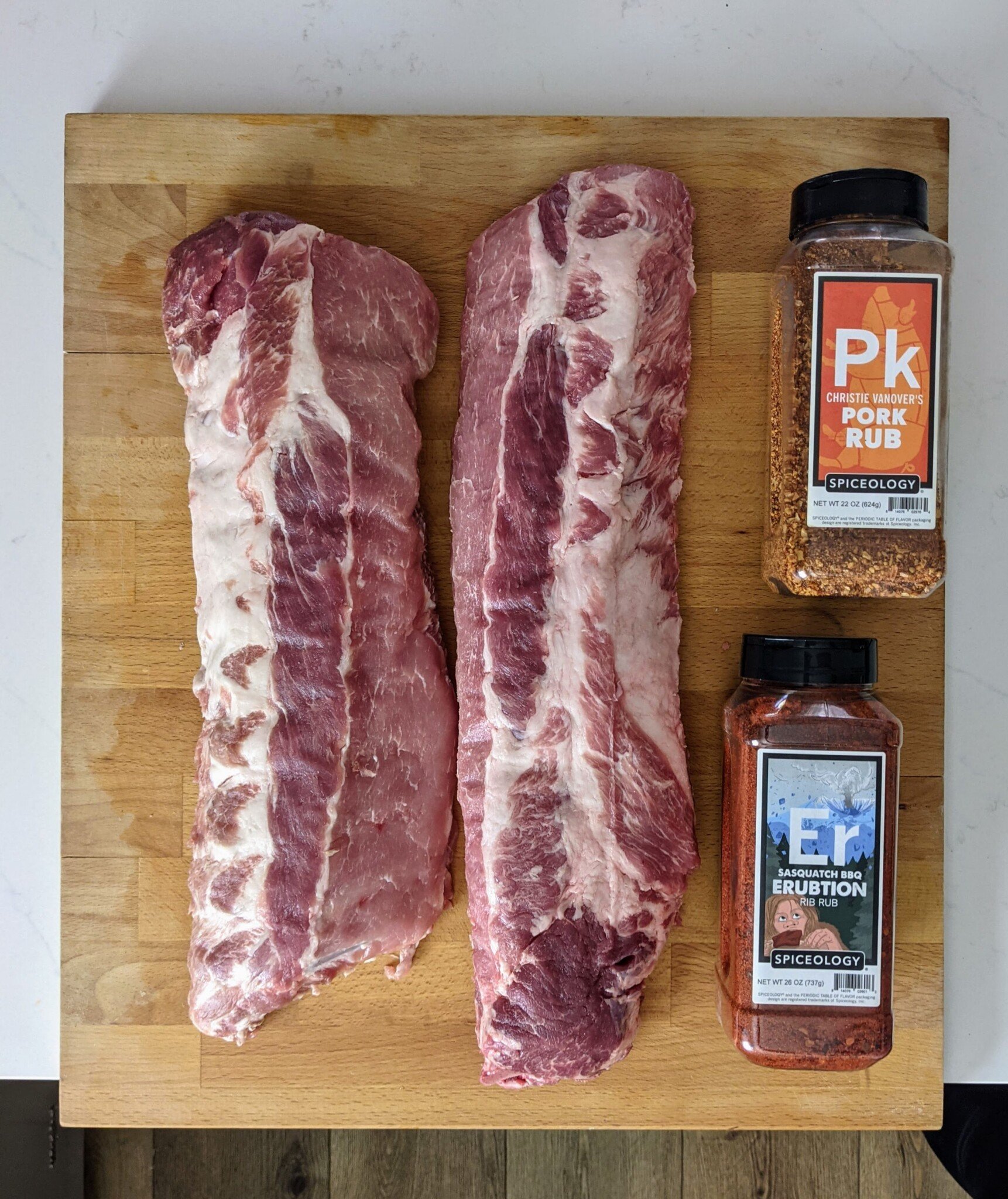 what you need to make easy dry rubbed smoked baby back rubs - showing the spice blends, PK and Erubtion with ribs on a cutting board 
