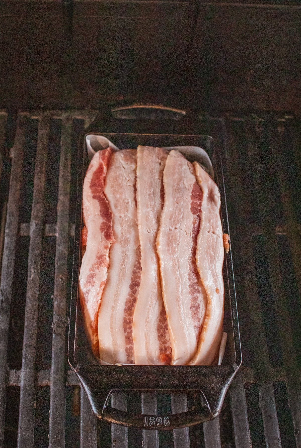 Bacon wrapped meatloaf on the grill grates, showing the bacon just starting to render, glistening.