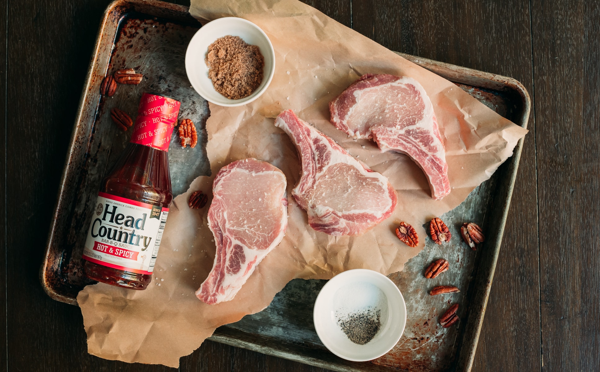 Overhead of raw pork chops on baking sheet with salt pepper, bbq sauce and pecans as additional ingredients. 