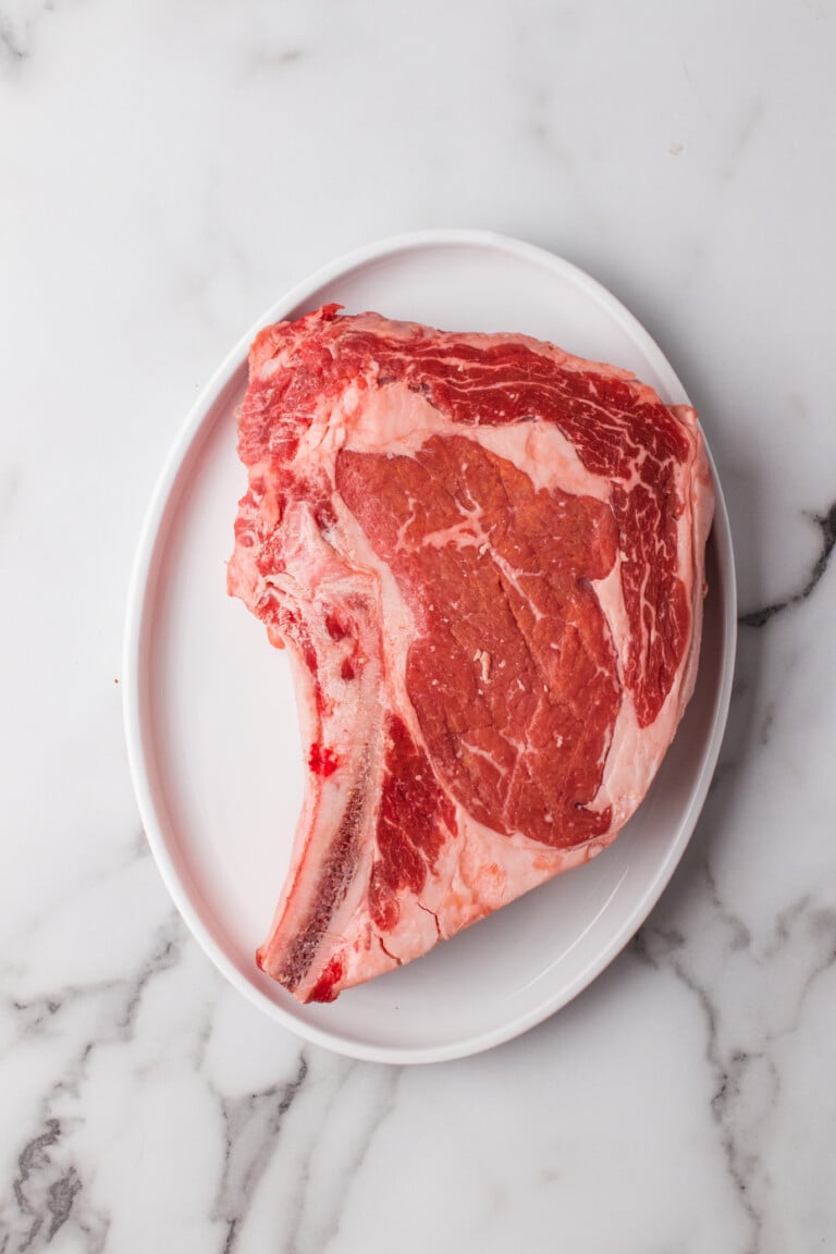 A raw bone-in ribeye on a white oval plate on marble, showing its marbling and fat cap.