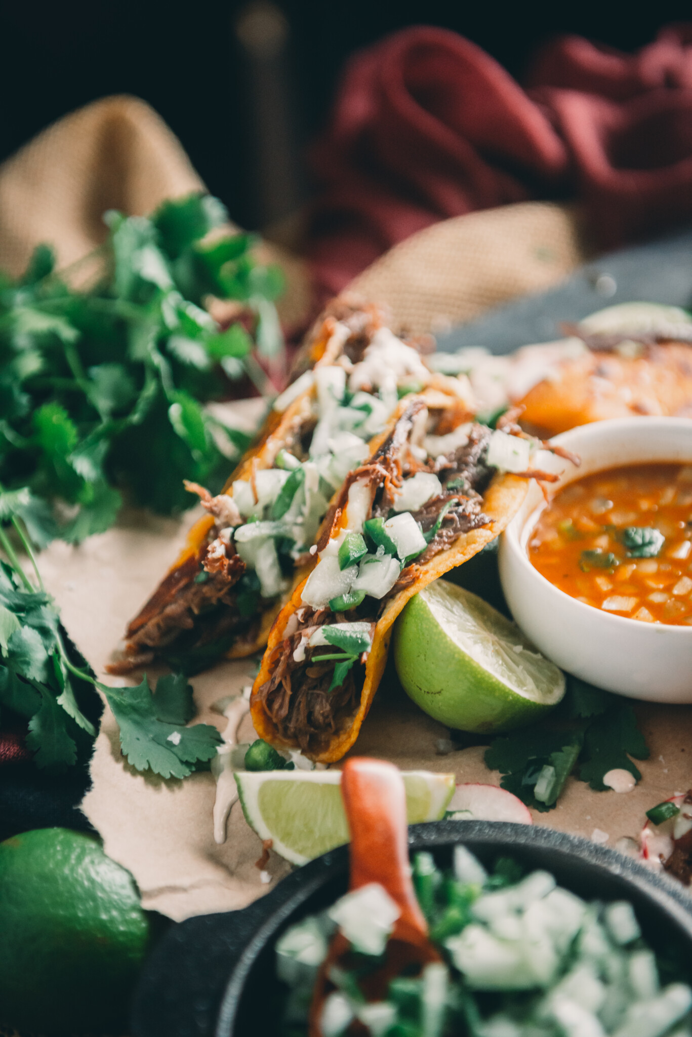 Bacllit image of limes, birria tacos and birria broth for dipping. 