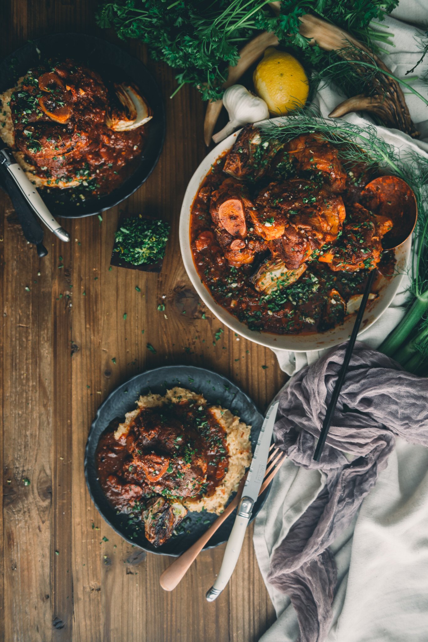 ovehead shot of table with platter of osso buco in sauce and black plates with the pork shanks, sauce, roasted fennel and mashed parsnips served 