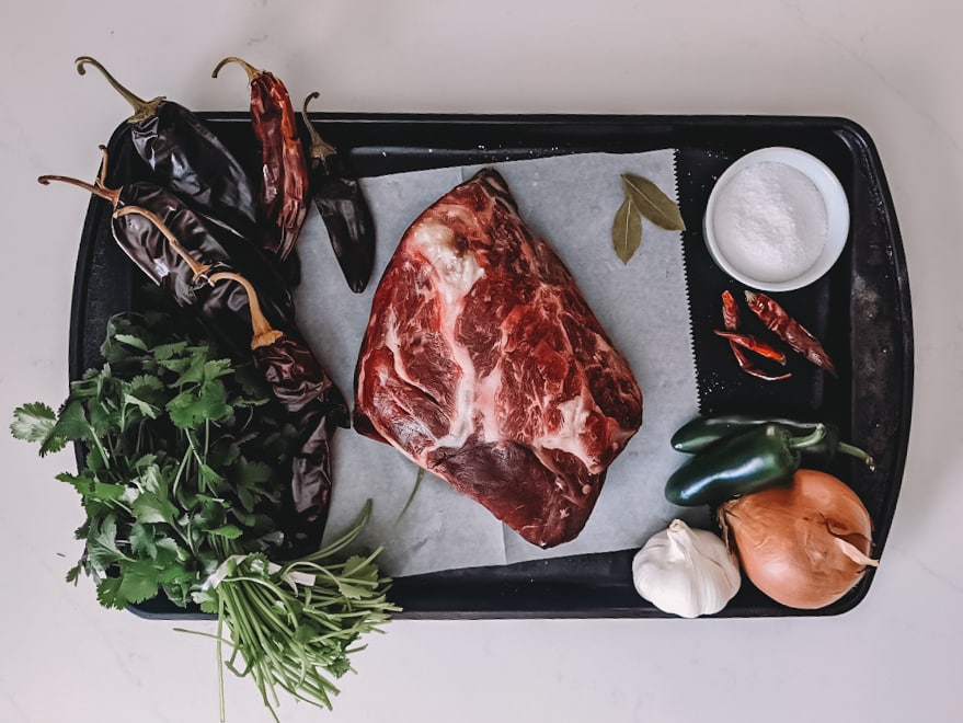 above shot showing ingredients for beef birria; beef neck, dried peppers, onion, garlic, jalapenos, bay leaves and salt