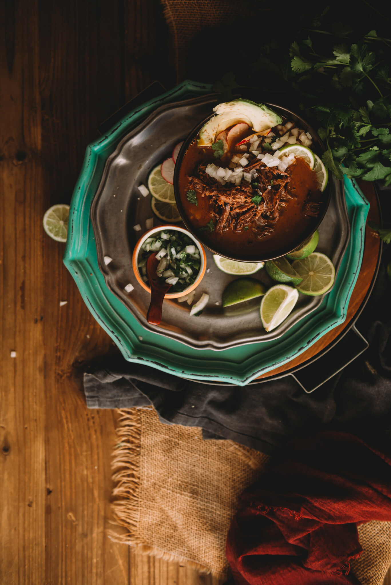 Overhead shot of birria in a bowl garnished with avocados, limes,  cilantro and onions. 