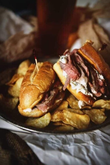 Platter with french dip sandwiches layered with sliced roast beef and onion dip with chips on the side.