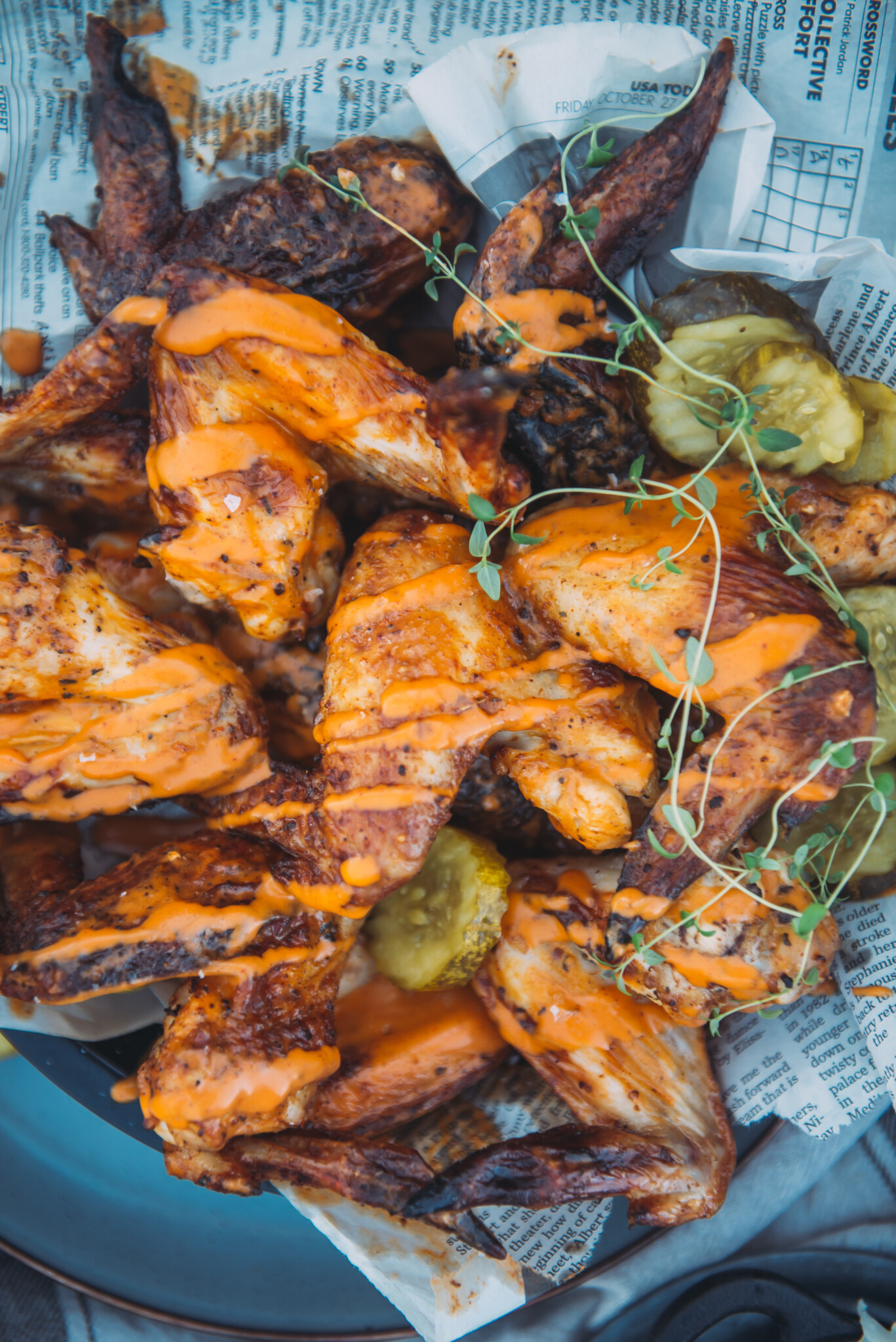 Close up of air fryer pickle brined chicken wings on a serving platter with pickles and thyme as garnish and a drizzle of buffalo sauce.