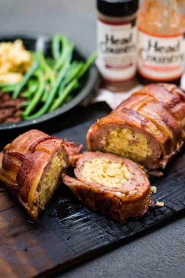 Bacon wrapped BBQ Fatty on a wooden slab with condiments and a plate of French beans and mashed potatoes behind it.