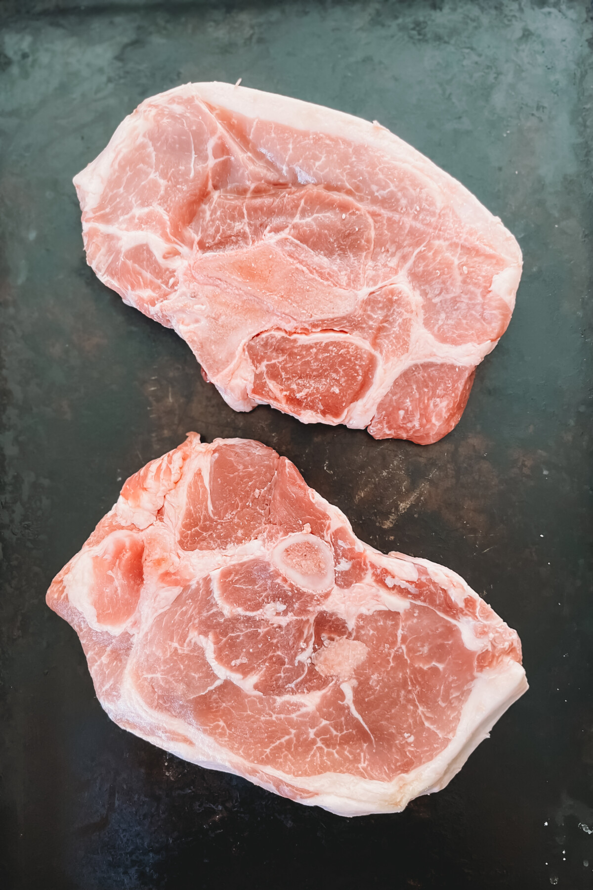 2 raw bone in porterhouse pork chops on black surface.