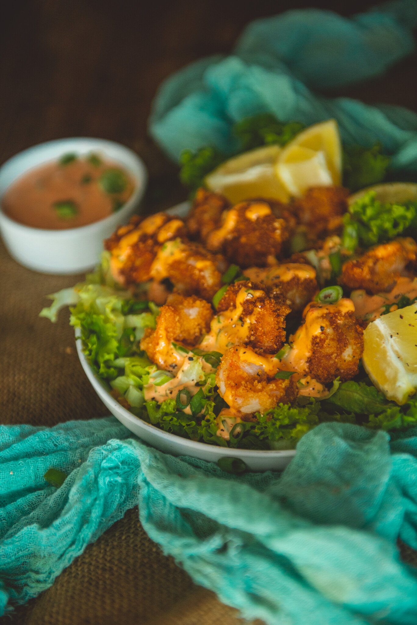 Close up shot of crispy pork panko crusted air fried shrimp in a bowl atop lettuce drizzed with bang bang sauce and garnished with lemon wedges. 