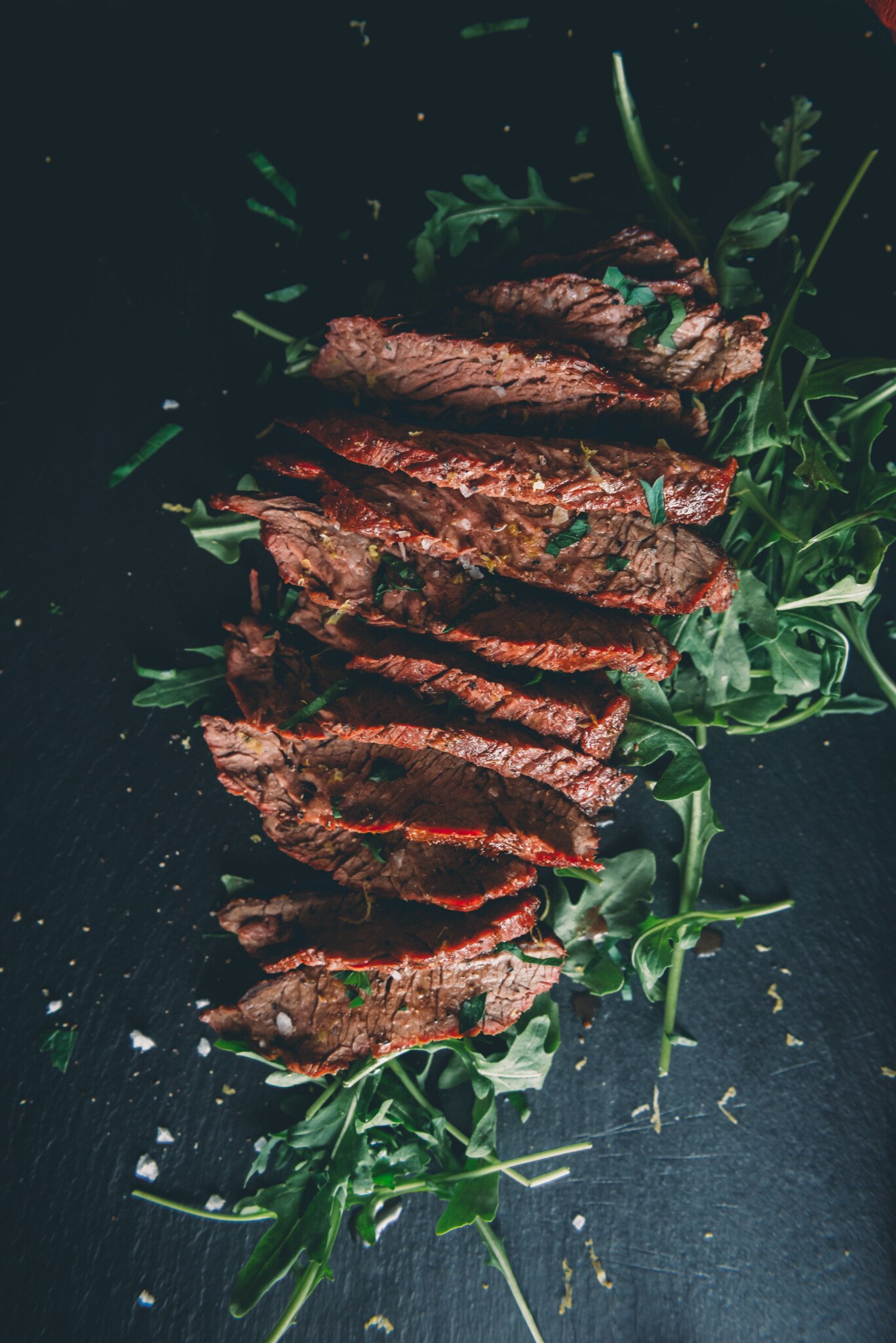 Above shot of sliced bavette steak that was grilled over charcoal arranged over a bed of green arugula and garnished with sprinkles of flakey salt. 