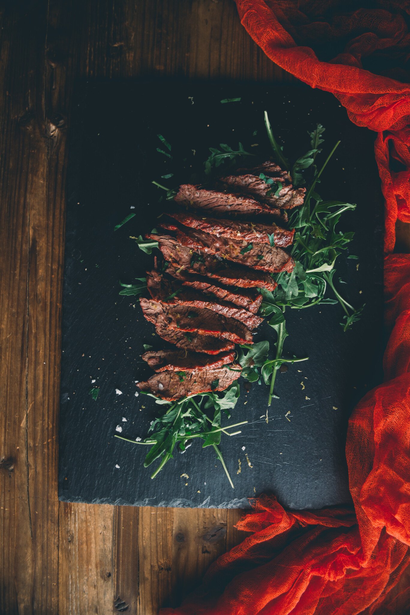 sliced char-grilled bavette steak sitting on a bed of arugula garnished with salt and snipped parsley on black slate. 