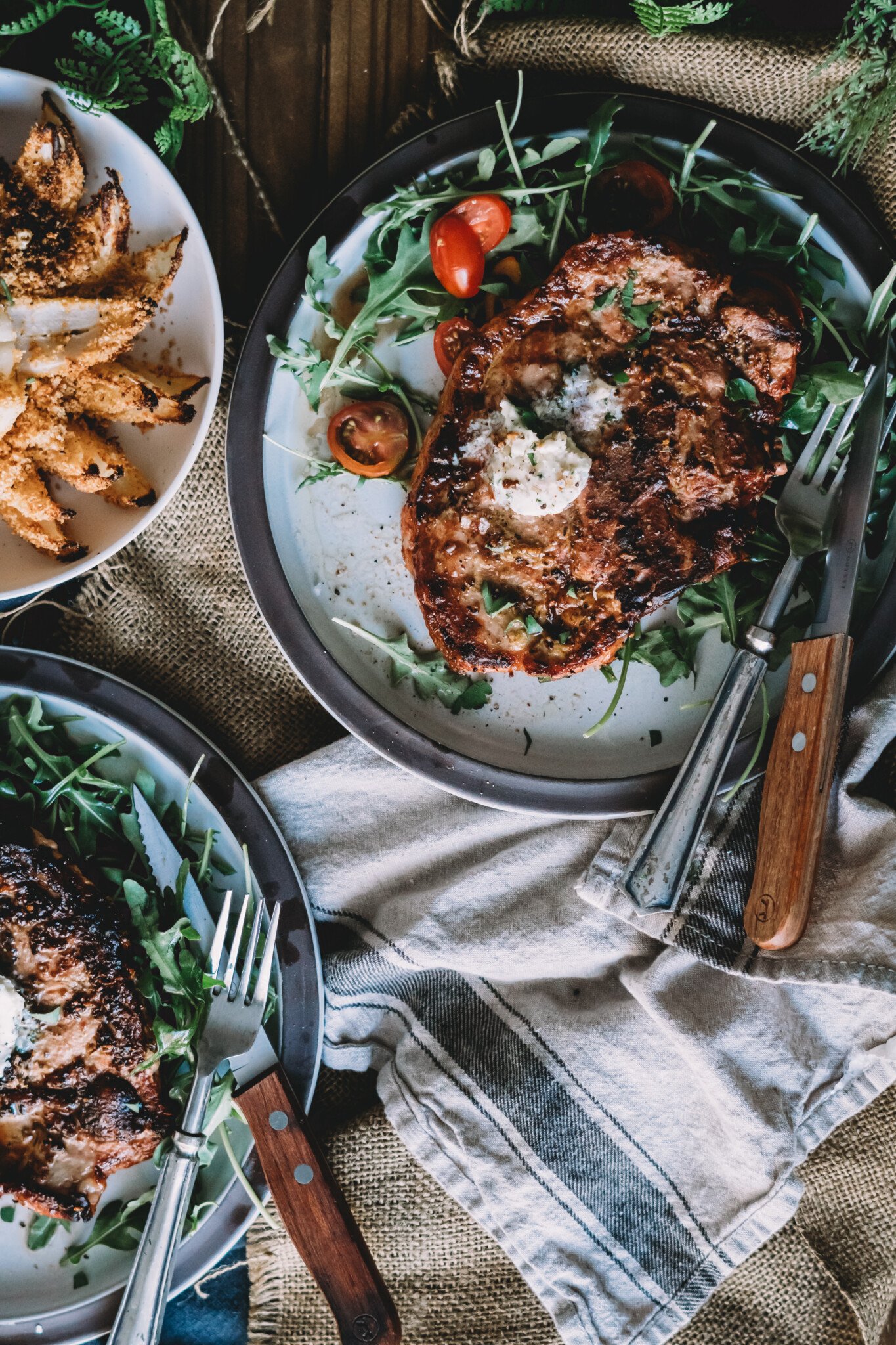 Above shot of 2 playes with grilled pork chops topped with compound butter over tossed arugula and cherry tomatoes; knives forks, and linens also dress the table.