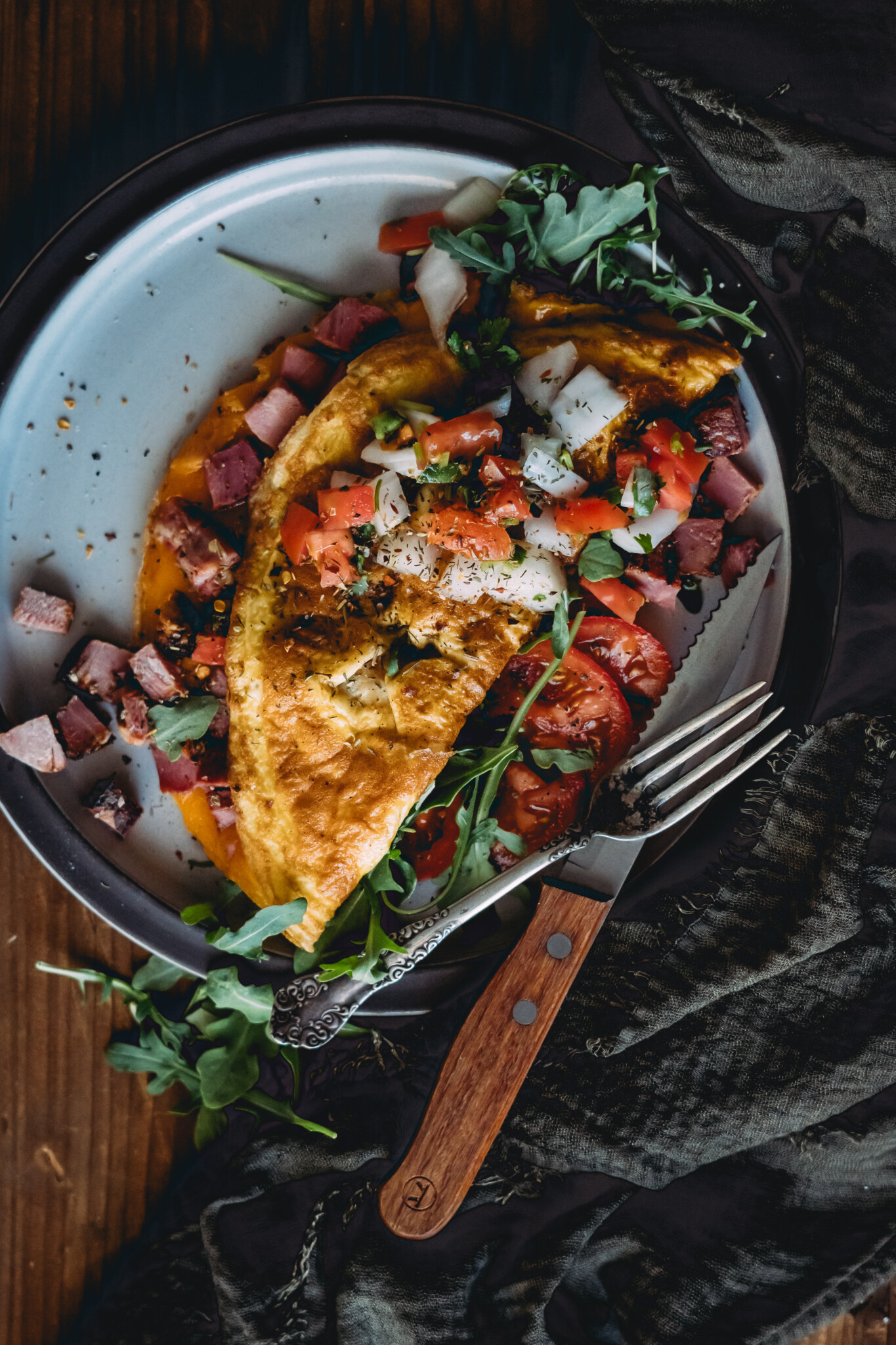 Large omelet on serving plate with diced ham falling out and topped with herbs and pico de gallo, atop arugula, knife and fork to the side. 
