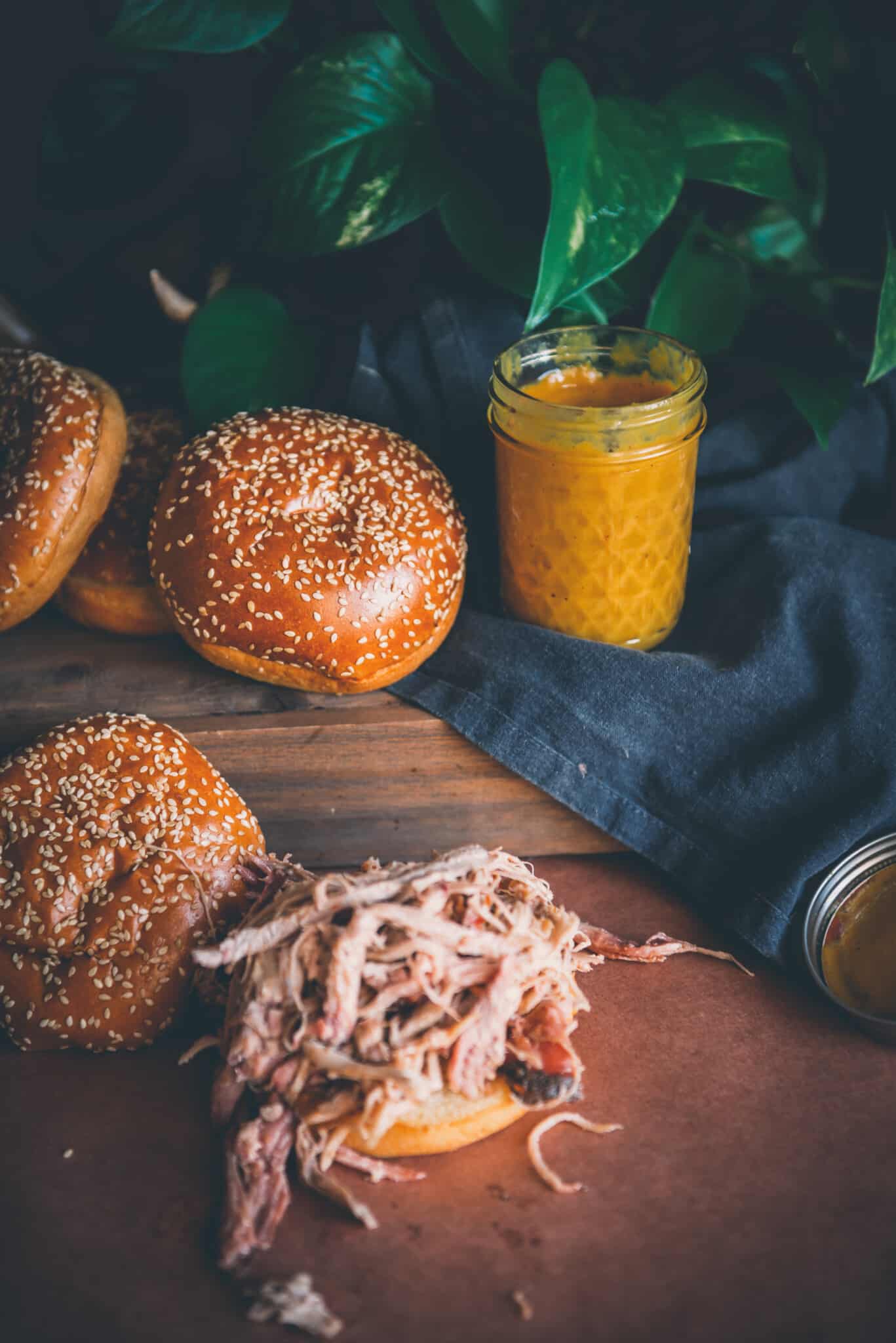 Jar of bright yellow mustard barbecue sauce on black linen near buns and pulled pork sandwich. 