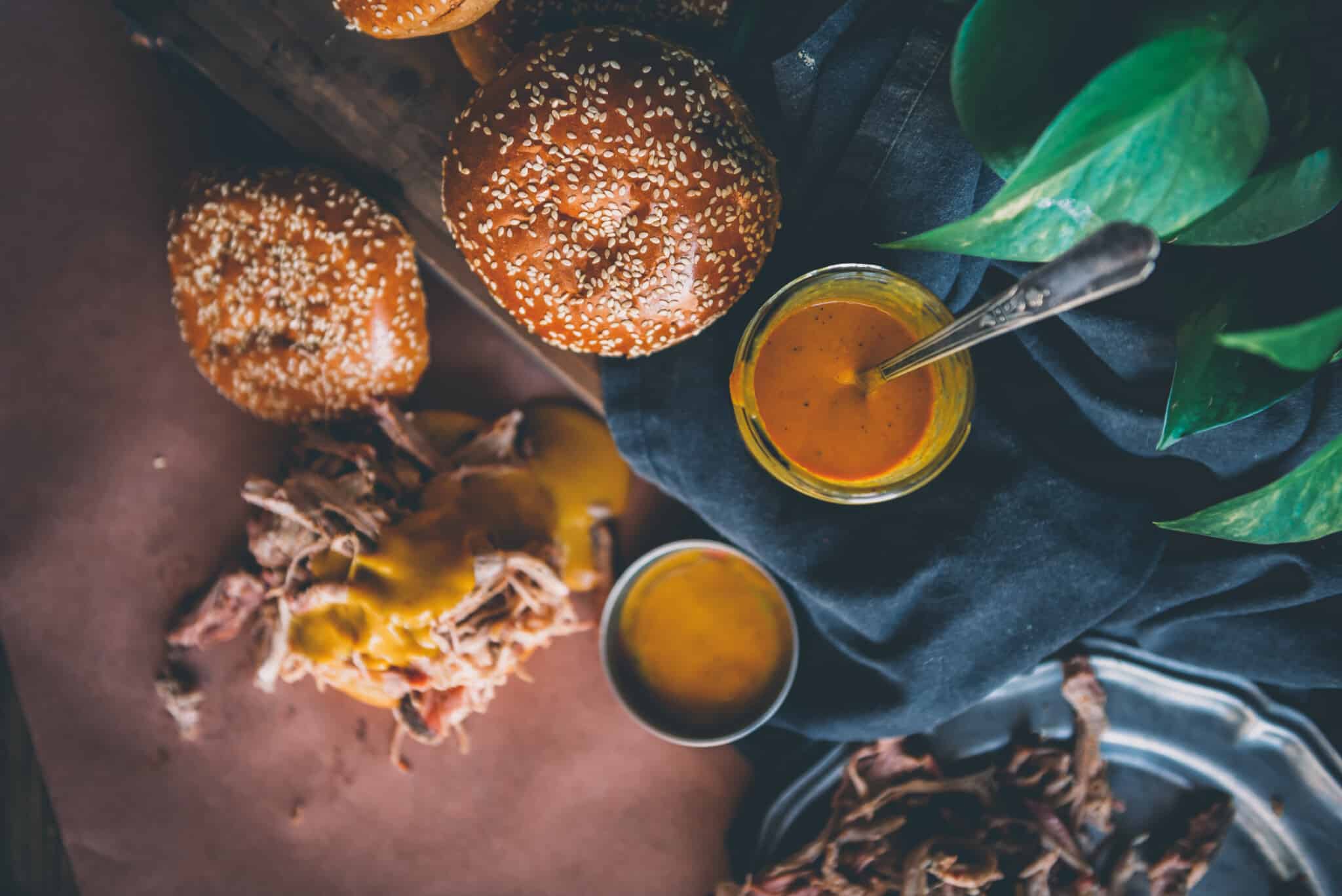 Over head shot of jar of mustard based bbq sauce with buns and pulled pork piled near by. 