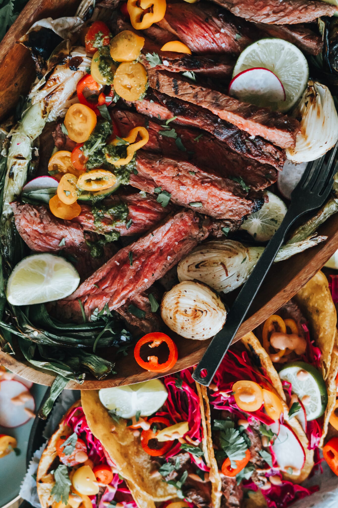 Close up of medium-rare grilled bavette steak sliced and on a platter with grilled spring onions and other taco toppings. 