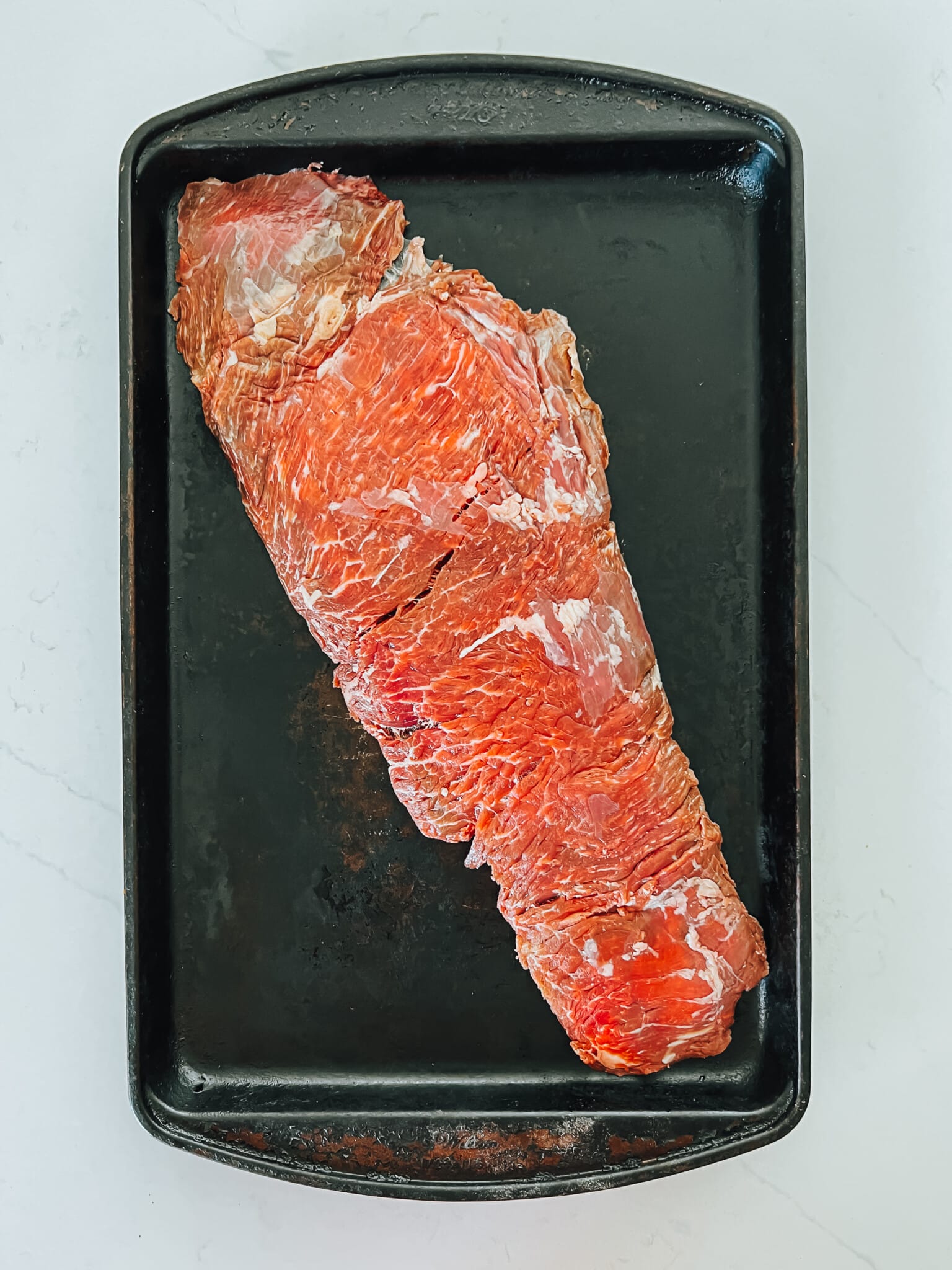 Whole bavette steak laid on to black baking sheet showing thin grain and marbling.  