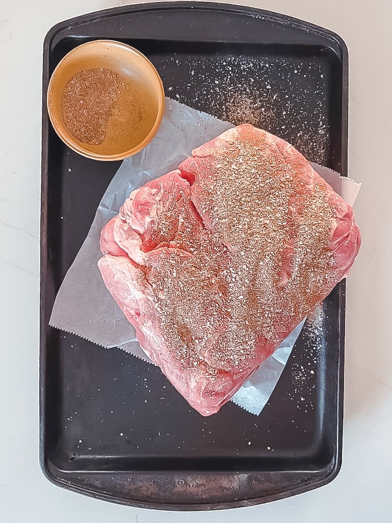 Raw pork butt on a sheet tray, seasoned with dry rub before smoking.