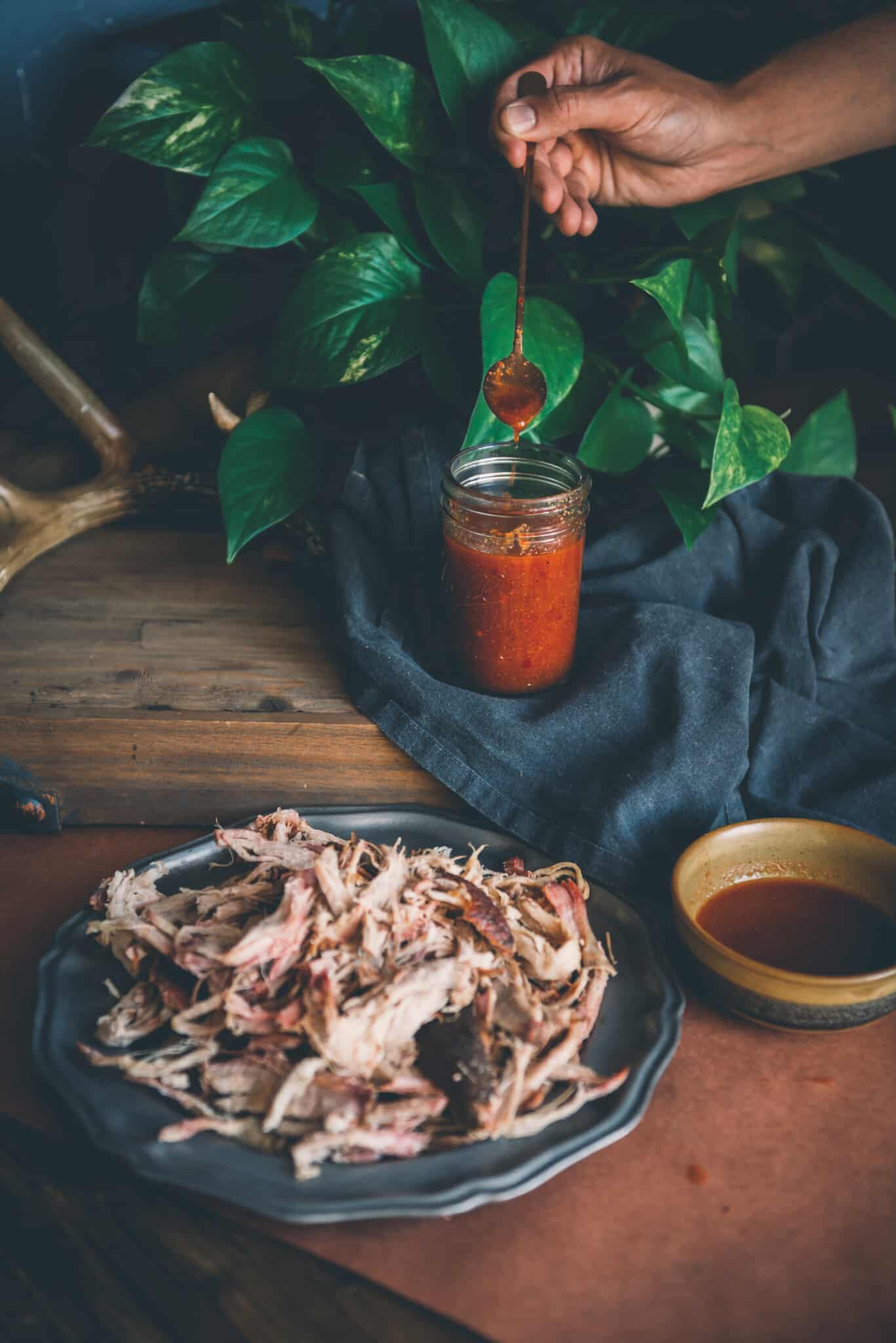 Hand picking up spoon of homemade vinegar bbq sauce our of a jar behind  platter of pulled pork.