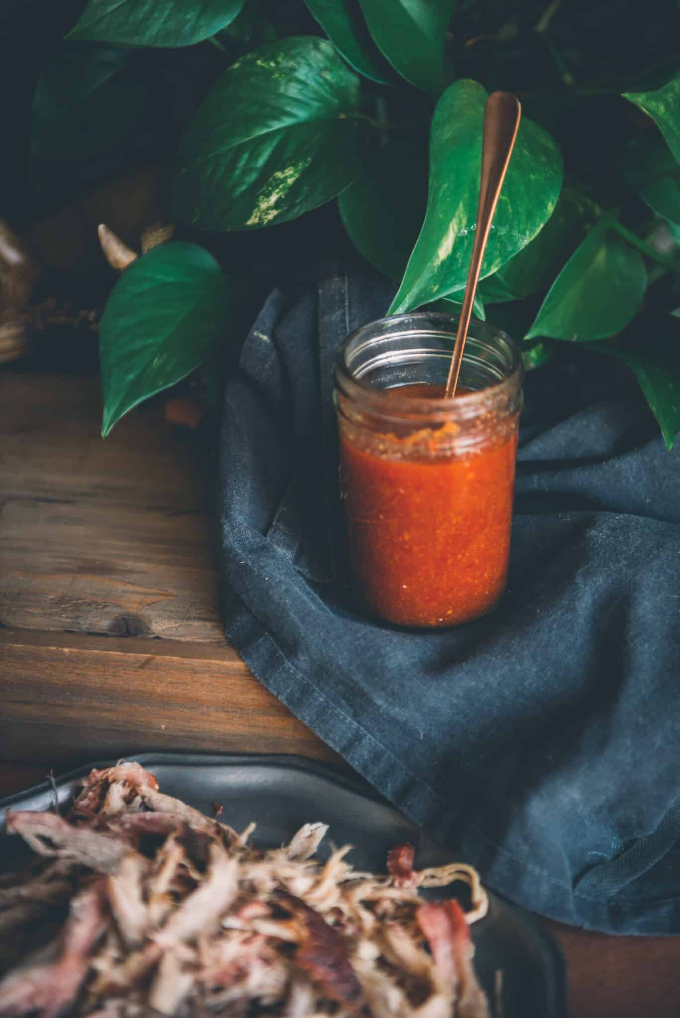 Open mason jar of vinegar based bbq sauce on black cloth with platter pf shredded pork in foreground.