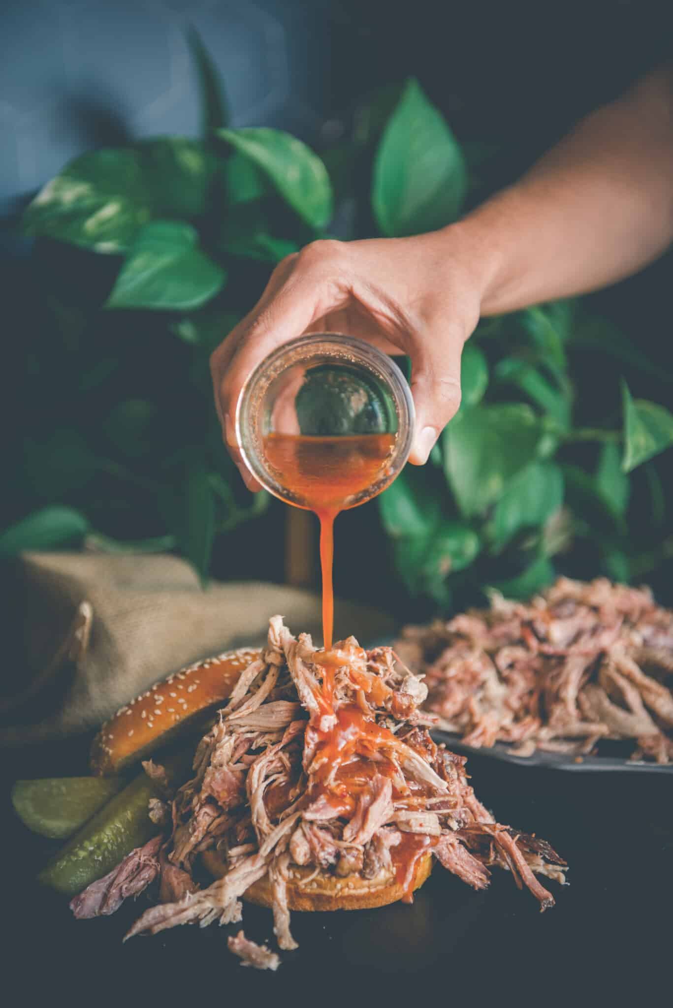 Hand pouring vinegar based sauce over pulled pork sandwich.