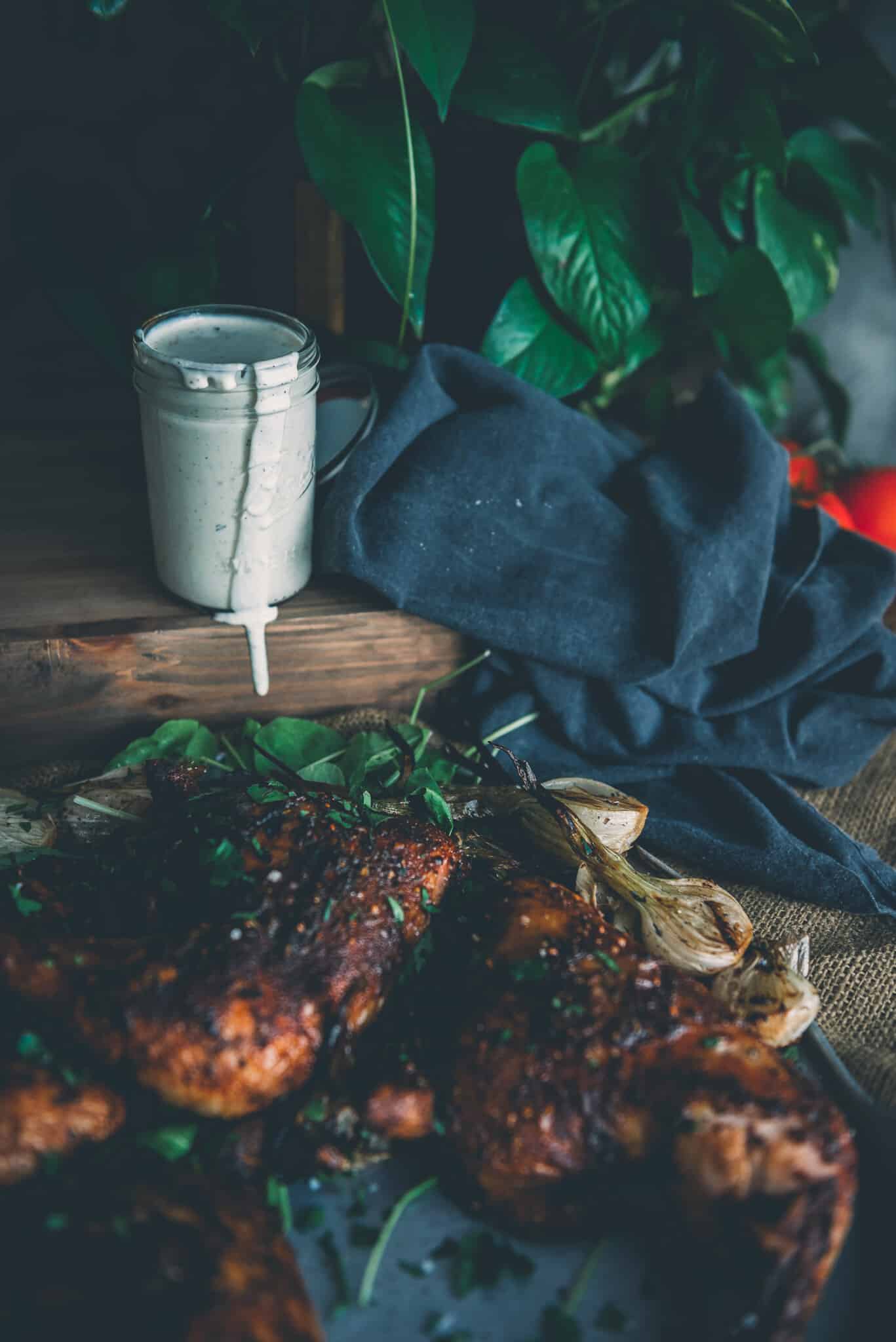 White Alabama BBQ Sauce in a jar with drizzle running down the side, on a wooden board, with chicken in the foreground. 